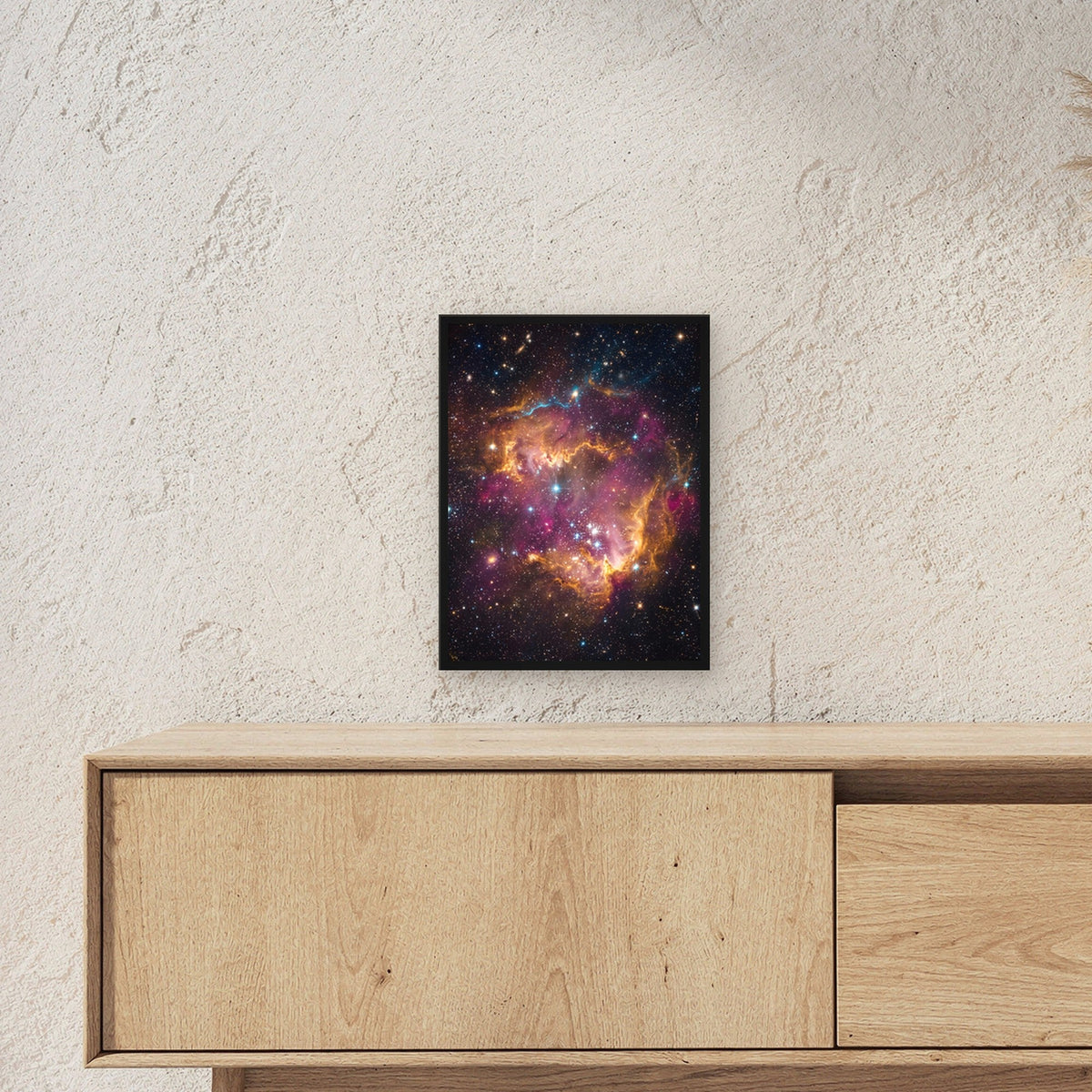 Die Sternenwiege – Sternennebel in Orange und Magenta mit jungen Sternen, im schwarzen Holzrahmen, über einem Sideboard